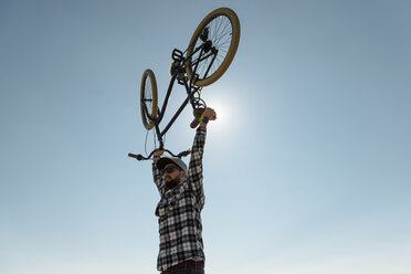 Niedriger Blickwinkel eines Mannes mit erhobenen Armen, der ein Fahrrad trägt, während er vor einem klaren blauen Himmel an einem sonnigen Tag steht - CAVF61088