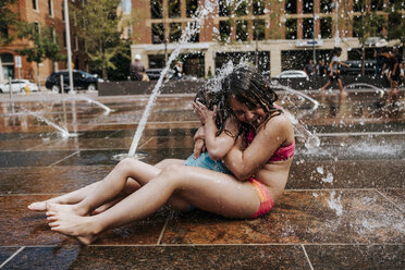 Seitenansicht von glücklichen Schwestern in Badekleidung, die mit Springbrunnenwasser vor Gebäuden in der Stadt spielen - CAVF61126