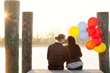 Rear view of couple with balloons kissing while sitting on pier - CAVF61153