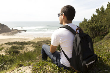 Seitenansicht eines Mannes mit Rucksack, der auf einem Hügel sitzt und auf das Meer schaut, gegen den Himmel an einem sonnigen Tag - CAVF61159