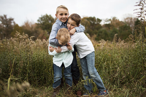 Porträt eines Jungen, der seine Brüder umarmt, während er auf einem grasbewachsenen Feld im Wald steht - CAVF61190