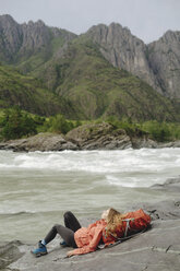 Female hiker reclining on backpack at riverbank against mountain - CAVF61200