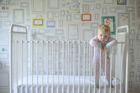 Porträt eines niedlichen kleinen Mädchens, das sich an ein Geländer lehnt, während es in einem Kinderbett an der Wand steht, lizenzfreies Stockfoto