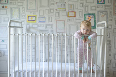 Porträt eines niedlichen kleinen Mädchens, das sich an ein Geländer lehnt, während es in einem Kinderbett an der Wand steht - CAVF61218