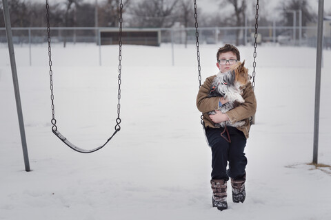 Porträt eines Jungen mit Hund auf einer Schaukel auf einem Spielplatz im Winter, lizenzfreies Stockfoto