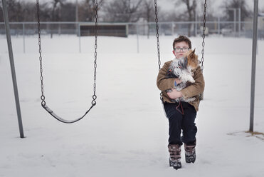 Porträt eines Jungen mit Hund auf einer Schaukel auf einem Spielplatz im Winter - CAVF61224