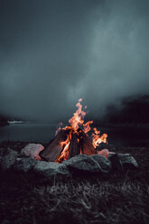 Lagerfeuer auf einer Wiese am Seeufer bei bewölktem Himmel in der Nacht - CAVF61243