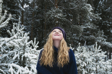 Happy woman with blond hair looking up while standing against trees in forest during winter - CAVF61342