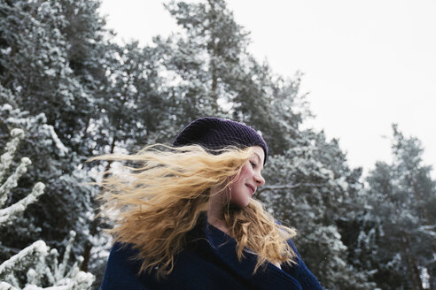 Niedriger Blickwinkel auf eine glückliche Frau mit blondem Haar, die wegschaut, während sie im Winter gegen Bäume im Wald steht - CAVF61343