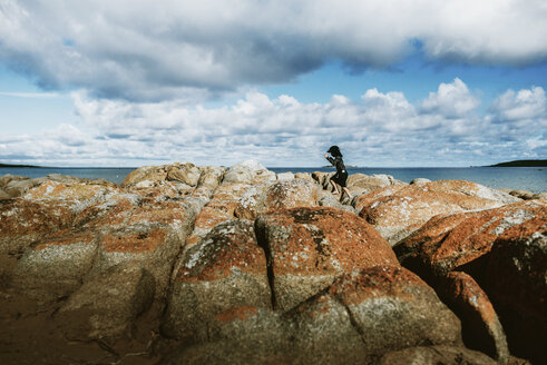 Seitenansicht von Frau springt auf Felsen am Strand gegen bewölkten Himmel - CAVF61397