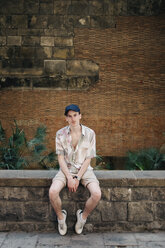 Portrait of confident man wearing cap while sitting on retaining wall - CAVF61401