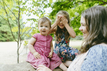 Glückliche Mutter steht bei Töchtern, die auf einem Baumstamm im Wald sitzen - CAVF61432