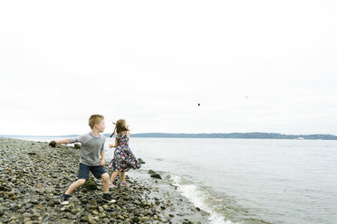 Playful siblings skimming stones in sea while standing at beach against clear sky - CAVF61438