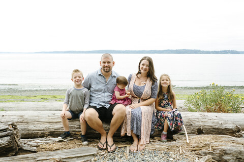 Porträt einer glücklichen Familie, die auf einem Baumstamm am Strand sitzt, gegen den klaren Himmel, lizenzfreies Stockfoto