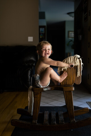 Seitenansicht eines fröhlichen kleinen Mädchens, das zu Hause auf einem hölzernen Schaukelpferd sitzt, lizenzfreies Stockfoto