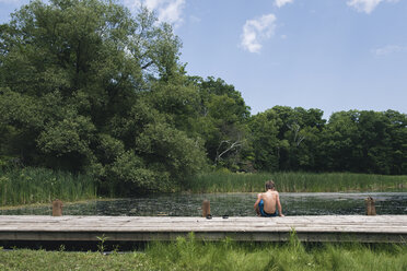 Rückansicht eines Jungen ohne Hemd, der auf einem Steg über einem See gegen den Himmel im Wald sitzt - CAVF61473