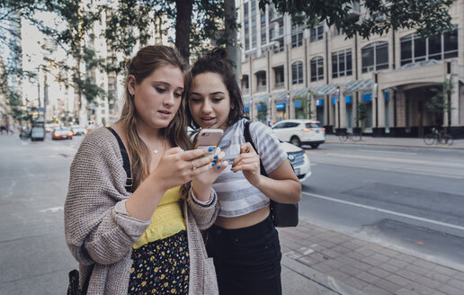 Freundinnen benutzen ein Smartphone, während sie auf dem Bürgersteig in der Stadt stehen - CAVF61483