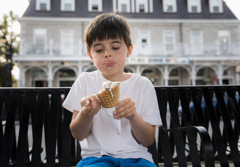 Süßer Junge isst Eiswaffel, während er auf einer Bank vor einem Gebäude in der Stadt sitzt, lizenzfreies Stockfoto