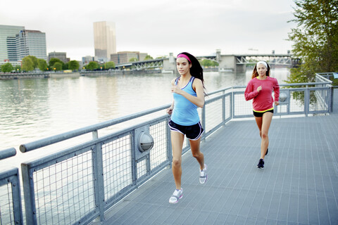 Weiblicher Athlet Freunde laufen auf Fußgängerbrücke über den Fluss gegen klaren Himmel in der Stadt, lizenzfreies Stockfoto