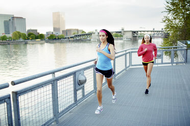 Female athlete friends running on footbridge over river against clear sky in city - CAVF61535