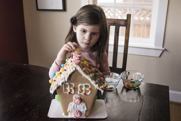 Cute girl decorating gingerbread house on table at home - CAVF61560