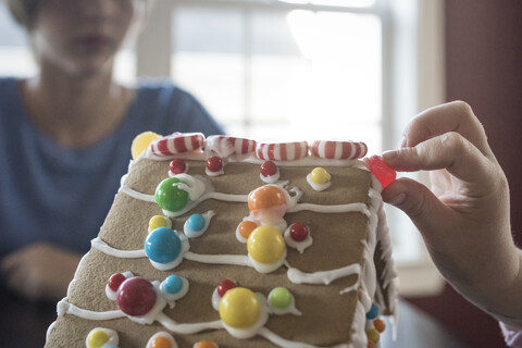 Abgeschnittene Hand eines Mädchens, das ein Lebkuchenhaus mit Süßigkeiten dekoriert, während die Schwester im Hintergrund zu Hause sitzt, lizenzfreies Stockfoto