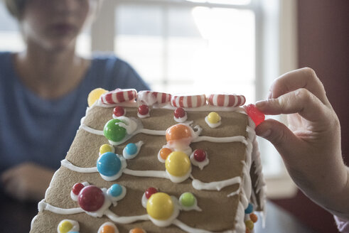 Abgeschnittene Hand eines Mädchens, das ein Lebkuchenhaus mit Süßigkeiten dekoriert, während die Schwester im Hintergrund zu Hause sitzt - CAVF61561