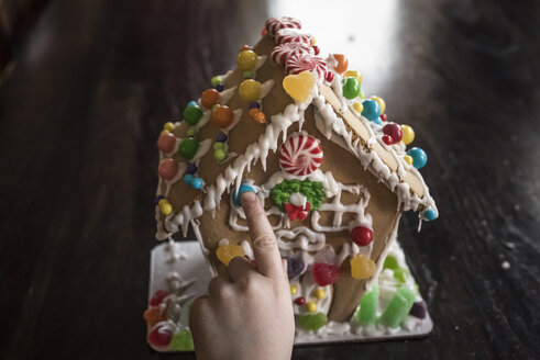 Abgeschnittene Hand eines Mädchens, das ein Pfefferkuchenhaus mit Süßigkeiten auf einem Tisch zu Hause dekoriert - CAVF61562