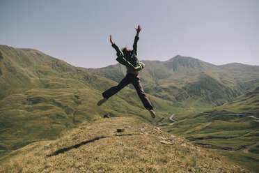 Rear view of happy woman with arms raised jumping on mountain against clear sky during sunny day - CAVF61610