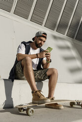Niedriger Blickwinkel eines selbstbewussten Mannes, der ein Mobiltelefon benutzt, während er mit einem Skateboard auf einer Stützmauer sitzt - CAVF61642