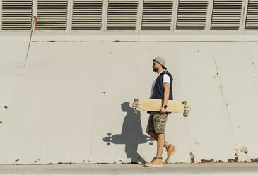 Seitenansicht eines Mannes, der ein Skateboard hält, während er an einem sonnigen Tag auf dem Fußweg gegen eine Mauer läuft - CAVF61644