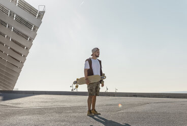 Selbstbewusster Mann, der ein Skateboard hält, während er auf der Straße steht, gegen einen klaren Himmel an einem sonnigen Tag - CAVF61646