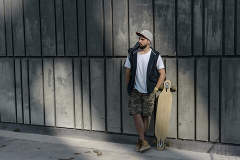 Nachdenklicher Mann mit Skateboard an einer Betonwand stehend, lizenzfreies Stockfoto