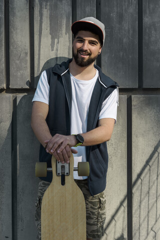 Porträt eines lächelnden Mannes, der ein Skateboard hält und an einer Betonwand steht, lizenzfreies Stockfoto