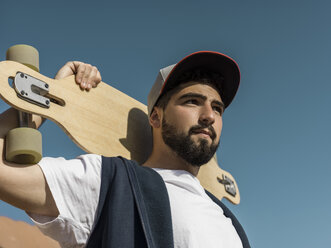 Low angle view of confident man holding skateboard while standing against clear blue sky during sunny day - CAVF61659