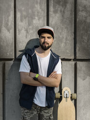 Porträt eines selbstbewussten, gut aussehenden Mannes mit verschränkten Armen, der mit einem Skateboard an einer Betonwand steht - CAVF61660
