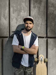 Portrait of confident handsome man with arms crossed standing by skateboard against concrete wall - CAVF61660