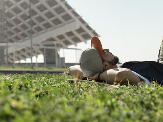 Surface level image of man with skateboard lying on grassy field against clear sky at park during sunny day - CAVF61665