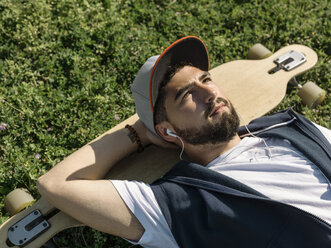 Hohe Winkel Ansicht der nachdenklichen Mann mit Skateboard Musik hören, während auf grasbewachsenen Feld im Park während des sonnigen Tages liegen - CAVF61667