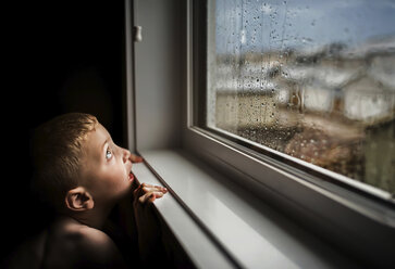Seitenansicht eines Jungen ohne Hemd, der während der Regenzeit zu Hause durch ein nasses Fenster schaut - CAVF61705
