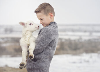 Seitenansicht eines niedlichen Jungen, der ein Lamm trägt, während er auf einem Bauernhof im Winter steht - CAVF61737