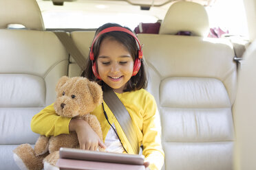 Lächelndes Mädchen mit Teddybär, das ein digitales Tablet mit Kopfhörern auf dem Rücksitz eines Autos benutzt - CAIF22799