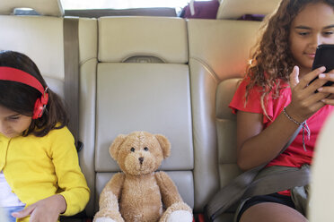 Schwestern mit Teddybär, die ein Smartphone und ein digitales Tablet benutzen und auf dem Rücksitz eines Autos sitzen - CAIF22822