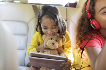 Niedliches Mädchen mit Teddybär, das ein digitales Tablet auf dem Rücksitz eines Autos benutzt - CAIF22824