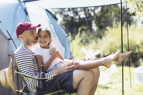Glücklicher, liebevoller Vater hält seine Tochter auf dem Schoß auf einem sonnigen Campingplatz - CAIF22834