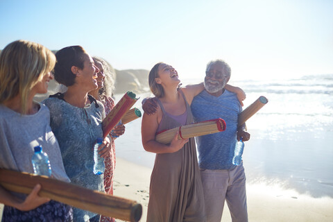 Glückliche Freunde mit Yogamatten, die sich am sonnigen Strand während eines Yoga-Retreats treffen, lizenzfreies Stockfoto