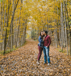 Man kissing happy woman while standing on dry leaves in forest during autumn - CAVF61750