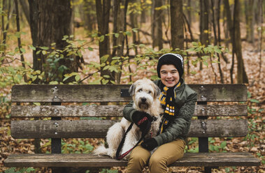 Porträt eines lächelnden Jungen mit Hund auf einer Bank im Wald im Herbst - CAVF61764