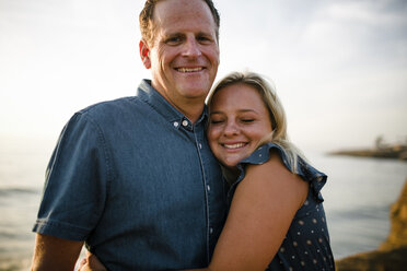 Porträt eines glücklichen Vaters, der von seiner Tochter umarmt wird, während er bei Sonnenuntergang auf einer Klippe am Meer steht - CAVF61792