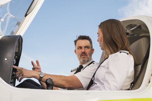 Niedriger Blickwinkel auf einen männlichen Piloten, der einem Flugschüler die Bedienung des Steuerpults in einem Flugzeug gegen den blauen Himmel am Flughafen beibringt - CAVF61798